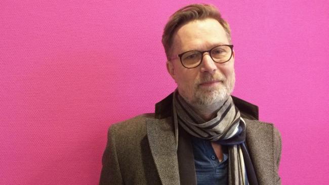 Porträtt Kenneth Nordgren med glasögon, rock och halsduk. Rosa bakgrund.
