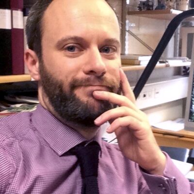 Selfie på Kent Fredholm med skägg, rödrutig skjorta och svart slips sittande på sitt kontor.