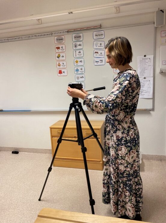 Kvinnlig lärare står vid en videokamera på stativ i ett klassrum.