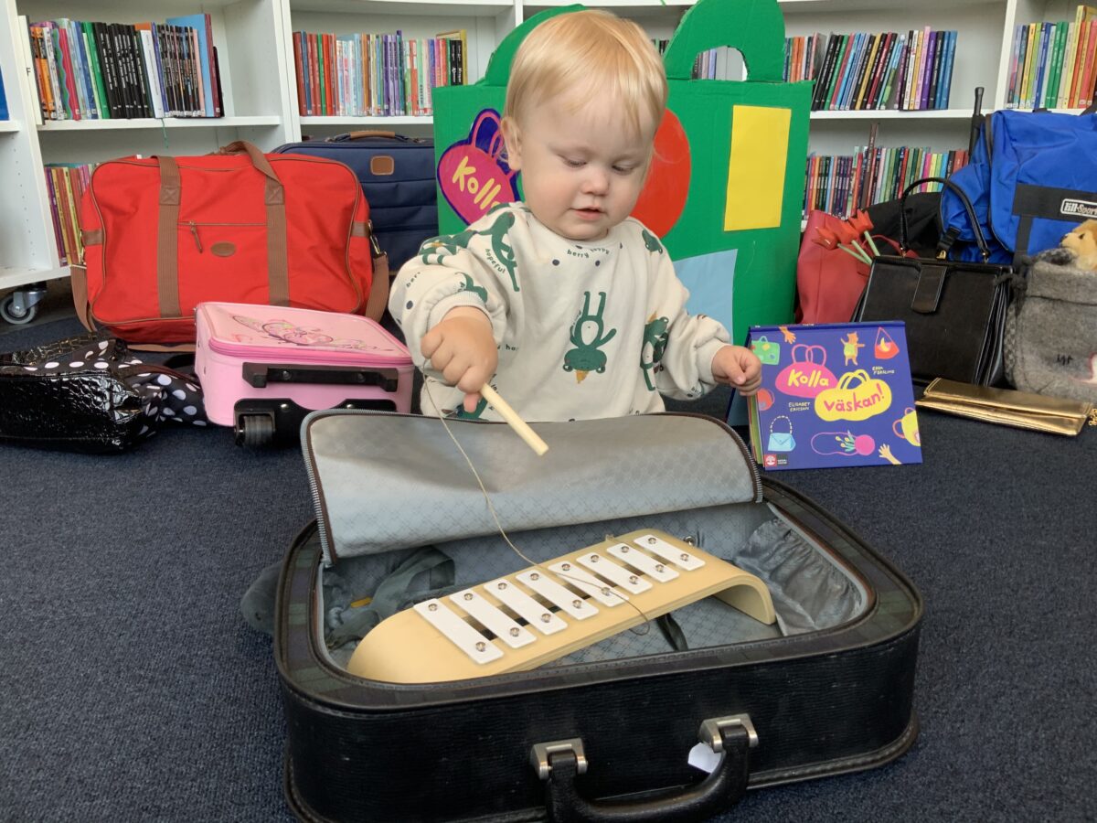 Ett barn på biblioteket spelar på en xylofon i en öppnad resväska. I bakgrunden en mängd väskor.