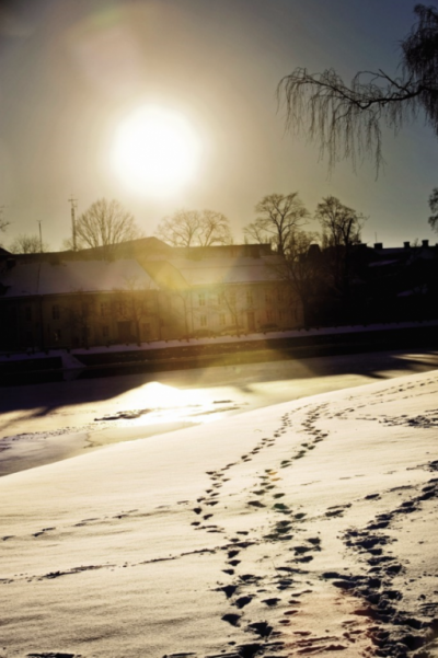Spår i snön fram ett fruset vatten och ett äldre hus. Solen går ner ovanför huset.