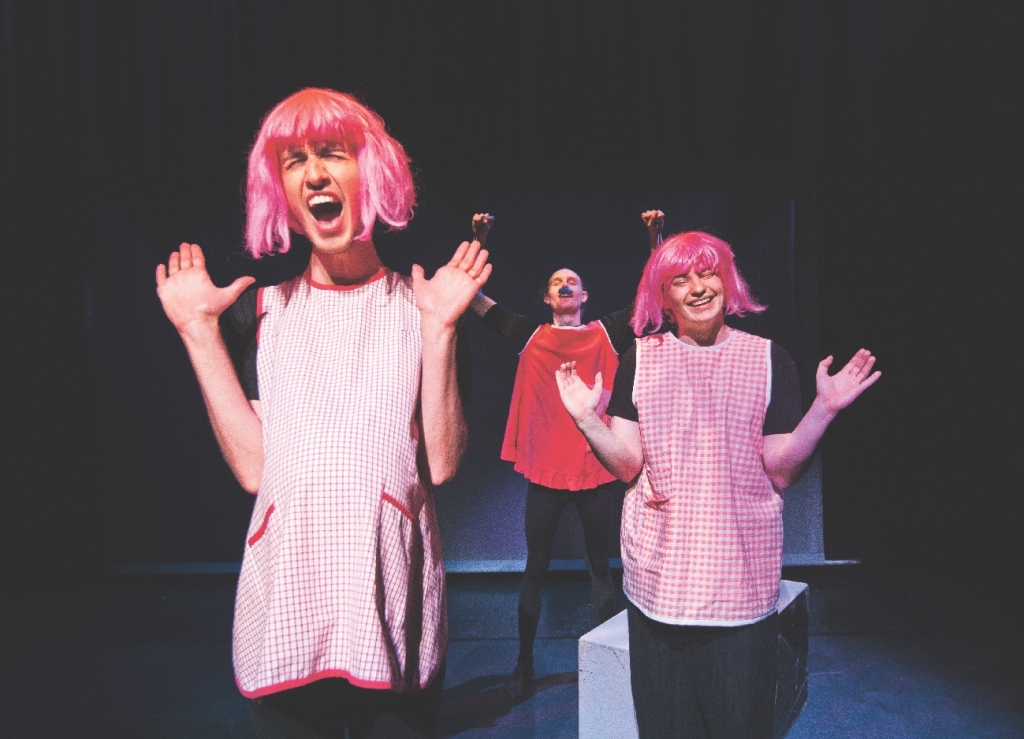 Två skådespelare med rosa peruker och rosa förkläden håller upp händerna. Bakom dem står en skådespelare i rött förkläde och mustasch.