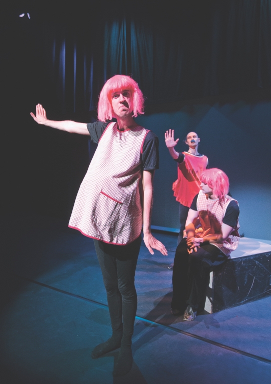 Tre skådespelare i rosa förkläden, två med rosa peruker. En sitter på en låda och två håller upp handen som för att visa "stopp".