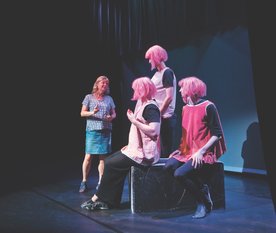 Teaterläraren Maria Hagberg Jangsell instruerar tre skådespelare med rosa förkläden och rosa peruk.