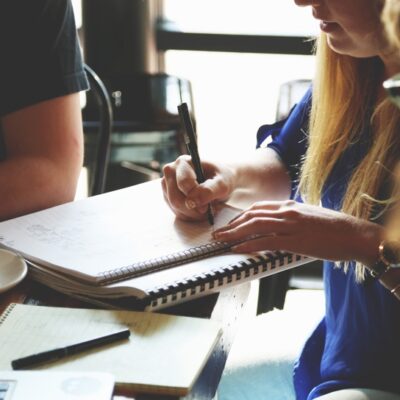 En person skriver i ett anteckningsblock med kollegor runt ett träbord. Det står en kaffekopp, laptop och skrivblock på bordet.