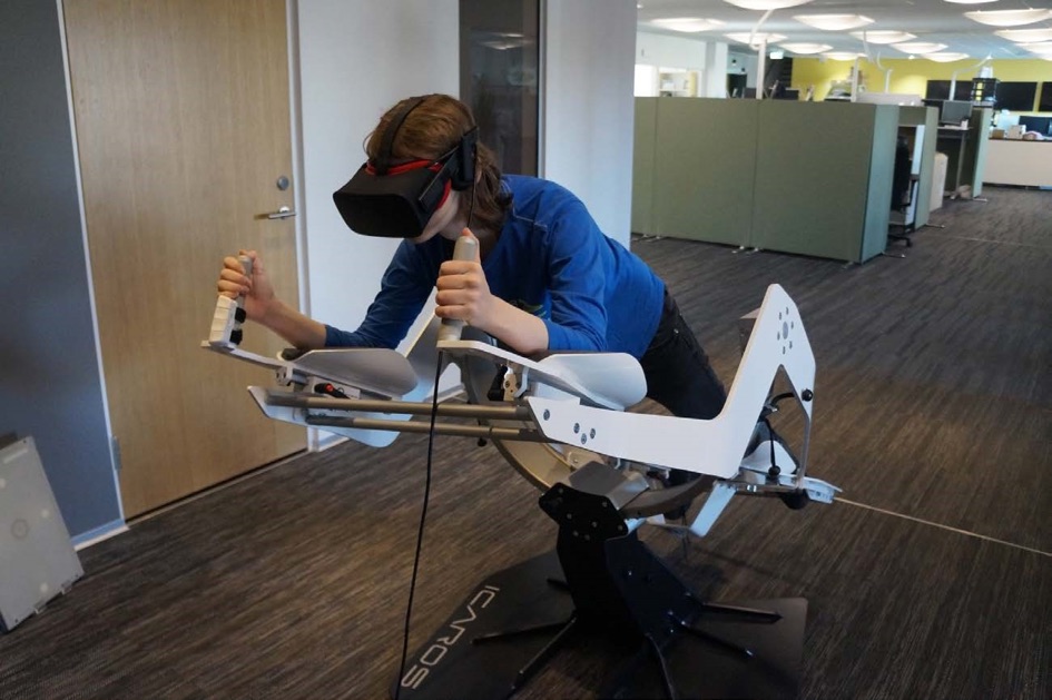 En ungdom i blå tröja ligger framåtlutad i en sorts flygsimulator med händerna på två spakar och VR-headset på huvudet.