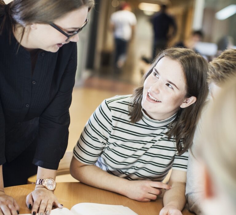 En lärare står bredvid och vänder sig mot elev som sitter.