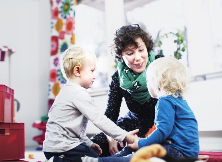 Två barn på förskolan sitter och håller varandra i handen och förskollärare sitter bredvid.